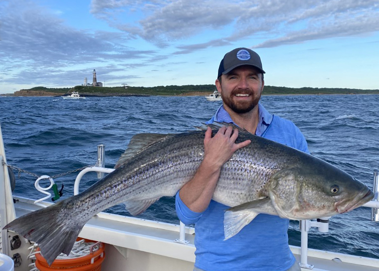 A man with the Montauk lighthouse behind him holding up a large sea bass