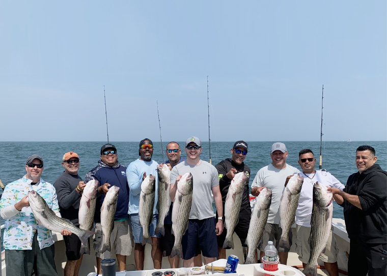 A group of men standing on the November Rain each holding a striped bass, with the ocean behind them