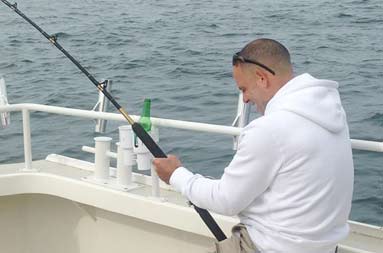 One of the guys sitting down on the boat reeling in a fish.