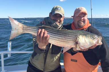Two men standing on the boat on a sunny day smiling into the camera as one of them holds up the bass he caught