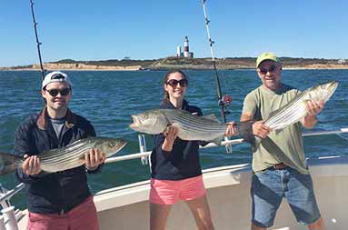A woman and two men hold up the striped bass they caught. The lighthouse is seen in the background.