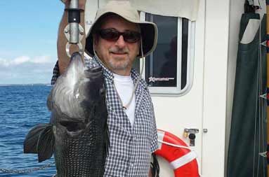A man wearing a beige Helios sun hat and sunglasses holds up a jumbo sea bass.