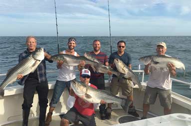 Six middle aged and senior men stand on the boat smiling, each holding a striped bass.