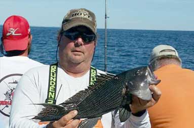 A man, wearing orange big pants and sunglasses, holds up a jumbo sea bass.
