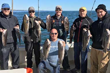 A group of six men aboard the boat dressed in winter attire holding up the blackfish they each caught.