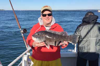 A man wearing a fleece and baseball cap standing in the sun holds up the blackfish (tautog) he caught.