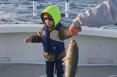 A bundled up young boy smiles as he gets some adult help holding up the striped bass he caught.