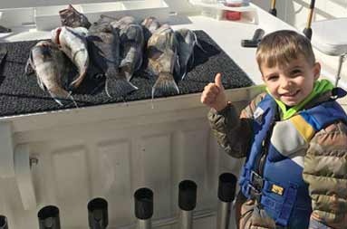 Still on the boat, back at the dock, a young boy smiles and gives a thumbs up standing next to a spread of striped bass on the fillet table.