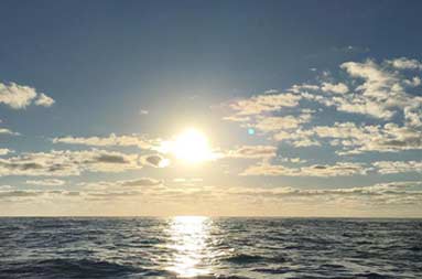 Dark waters and blue sky with a bright sun rising above it.