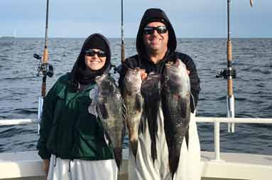 A woman and a man on the boat bundled up, each holding 2 jumbo sea bass.