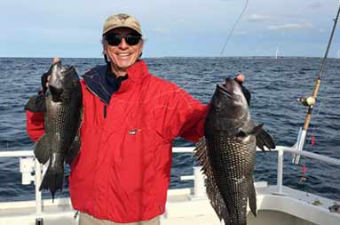 A man wearing a red jacket smiles as he holds up the two jumbo sea bass he caught.