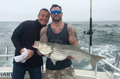 One guy proudly holds up the striped bass he caught while his friend stands next to him and smiles.