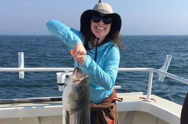 A woman smiles and proudly holds up the striped bass she caught.