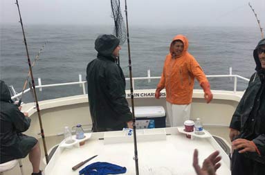 View from the cabin looking out at the deck on a cloudy, rainy day with Captain Steve in his orange raincoat and the group talking and fishing.