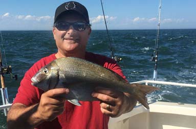 A man wearing a red t-shirt and sunglasses smiles and holds up the scup/porgy he caught.