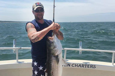 A guy wearing American flag board shorts smiles and holds up a large fish.