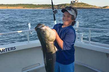 A young boy proudly holds up a large striped bass almost as long as himself.