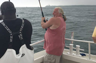 A man in the foreground on the boat smiling as he reels in his catch. First mate, Gil, watches in the background.