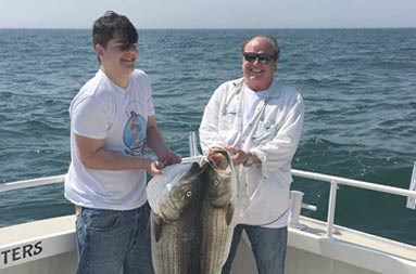 A man and younger man each smile proudly and hold up their catch of striped bass. 