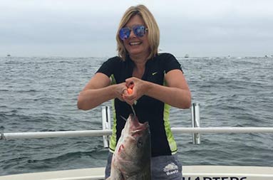 A woman wearing a black t-shirt holds up a striped bass by the hook.