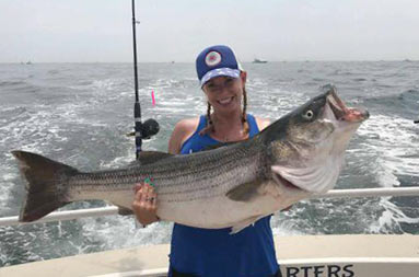 A woman wearing a blue baseball cap and blue tank top proudly holds up, with both hands, the large striped bass she caught .