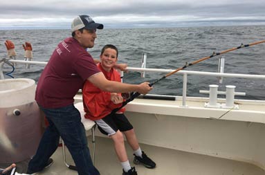An adolescent boy sits in the chair with his reel in the water as a man helps him reel the fish in.