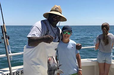 First Mate Gil puts his arm around a young boy as they both smile and hold up the sea bass he caught.