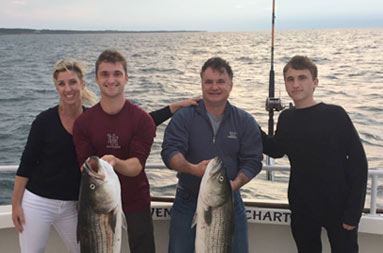 A mom and dad stand with their two teenage sons as one of the sons and father hold up striped bass.