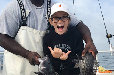 First mate, Gil, holds up the line with 2 sea bass, and the young boy who caught them has a very excited smile on his face as he points to the fish on either side of him with his thumbs.
