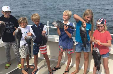 Six young/adolescent female and male children stand on the deck of the boat, all holding up the fish they caught.