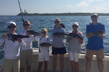 Two female adults, one male adults and 3 young adolescents each smile hold up their sea bass.