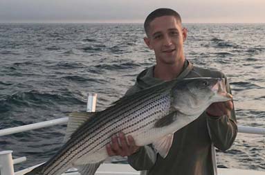 Just as the sun is rising behind him, a man holds up the striped bass he caught.