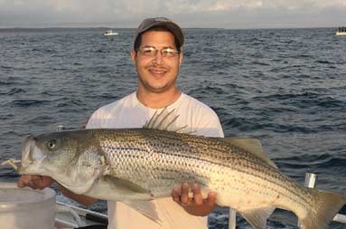 A man smiles as he holds the large striped bass he caught with the sun behind him still rising to daylight.