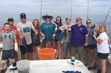 A group of 11 adults and young adults smile as some hold up the sea bass they caught.