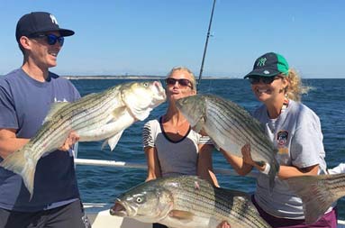 A woman and man each hold up their striped bass while another woman between them, holding up her own bass, pretends to kiss the fish.