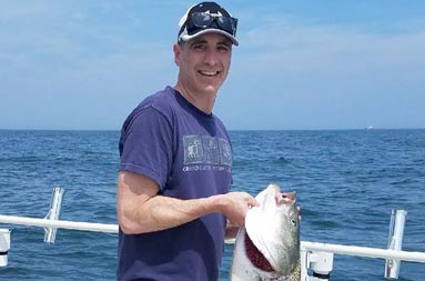 With the dark blue ocean behind him, a man holds up the striped bass he caught.