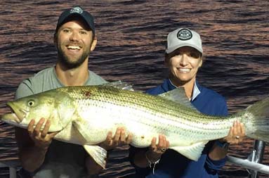 With the orange setting sun and lighthouse behind them, a man and woman smile as they hold up a bluefish.