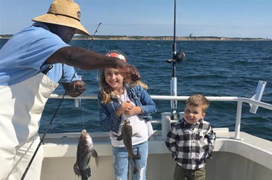 First mate, Gil, holds up two sea bass as the young girl and boy smile and stand next to their catch.