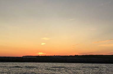View over the darkened water and silhouette of the land ahead with a blazing orange sunset in the background.