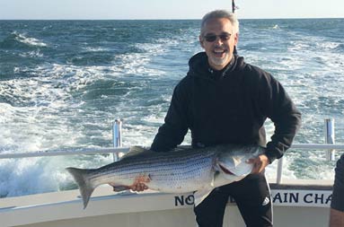 A man wearing sunglasses and black hoodie, smiles as he holds up his striped bass.