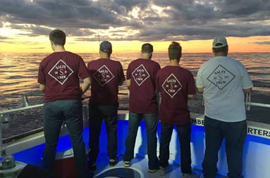 With a dark cloudy purplish sky above and a bright yellow/orange sunset in the distance, five men have their backs facing the camera to show off their celebratory t-shirts.
