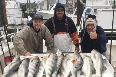 Three men wearing rain attire stand on the boat, by back at the marina, next to the fish they caught.