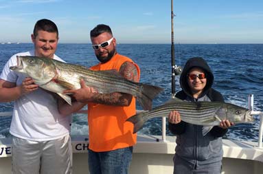 A man and teenage boy together hold up a striped bass, as a younger boy holds up a striped bass on his own.