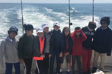 Nine girls and boys smile for the camera on the boat with the waves from the wake behind them
