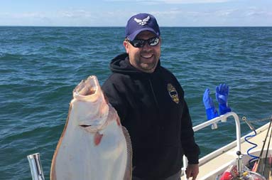 With the dark bluish/green ocean behind him, a man smiles big holding a large fluke with one hand.