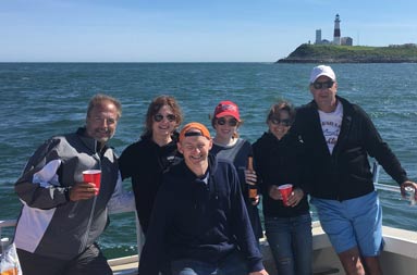 With the sun shining  on the water behind and the lighthouse in clear view, a family of six  smile for the camera.