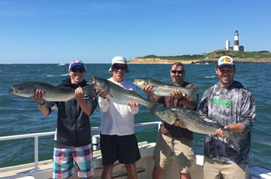 On a sunny day with the Montauk light house in the near distant background, 4 men smile and each hold up striped bass.