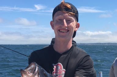 A young man with a bit smile holds up a large sea bass.