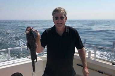 On a sunny day, a man holds up a sea bass and smiles.