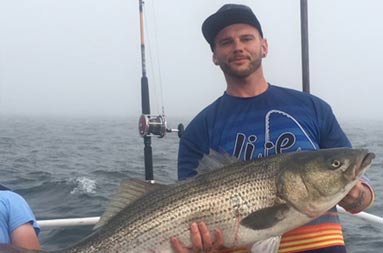 A man wearing a blue t-shirt and baseball cap holds up a large striped bass.